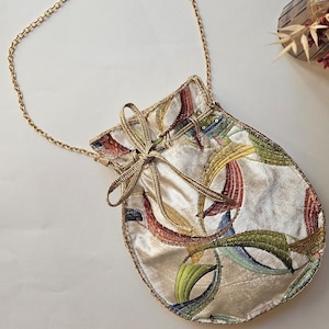 Könnte beinhalten: Eine weiße Samt-Kordeltasche mit einem goldenen Kettenriemen. Die Tasche hat ein farbenfrohes abstraktes Muster mit roten, gelben, grünen und blauen Akzenten.