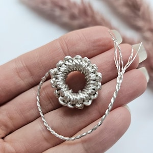 Op de afbeelding: Een zilverkleurig, gehaakt haaraccessoire met een cirkelvormig ontwerp en een gedraaid koord. Het accessoire wordt in een hand gehouden tegen een witte achtergrond. Het gehaakte stuk heeft een bloemachtig uiterlijk.