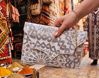 May include: A white clutch with a geometric pattern in gray and gold is held by a person. The clutch has a gold chain strap. The background includes colorful textiles and spices, suggesting a market setting.