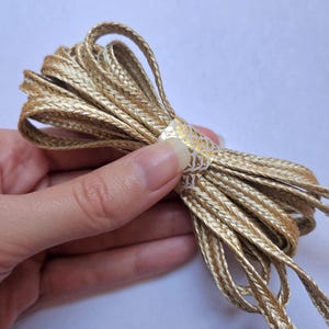 May include: A bundle of gold-colored braided cord, tightly wound and secured with a decorative band. The cord has a woven texture and a metallic sheen, suggesting its use in crafts or embellishments. The band features a white and gold pattern.
