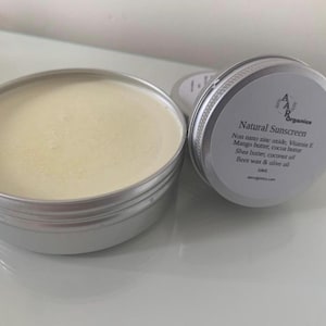 Pode incluir: Duas latas de prata abertas de protetor solar natural. Uma lata é preenchida com um creme amarelo claro. A outra lata, parcialmente visível, tem um rótulo que diz "Natural Sunscreen" com uma lista de ingredientes, incluindo óxido de zinco e manteiga de karité.
