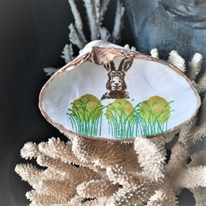 May include: A white seashell with a painted design of a rabbit in a field of green grass. The shell is sitting on a white coral.