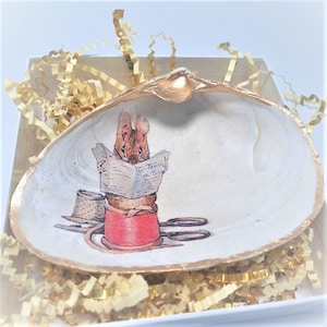 May include: A decorative seashell with a gold-painted rim, featuring a whimsical illustration of a rabbit reading a newspaper atop a spool of red thread. A small hat and scissors are also depicted. The shell is presented in a white box with gold filler.