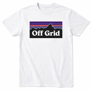 Op de afbeelding: Wit T-shirt met een rechthoekige afbeelding. De afbeelding toont een berglandsilhouet, horizontale strepen in paars, rood en blauw en de witte woorden "Off Grid" in een schreefloos lettertype. Het shirt heeft een ronde hals.