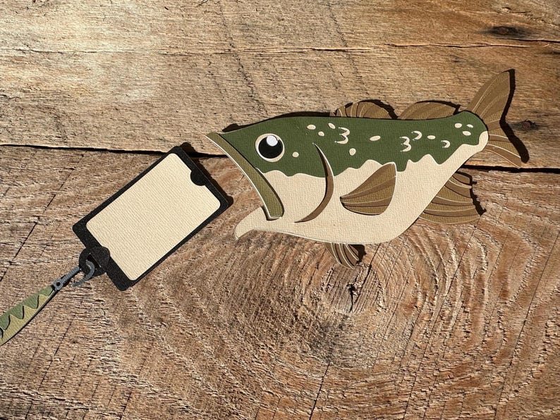 May include: A brown and green paper fish with a black and beige tag attached. The fish has a large mouth and a black eye. The tag is blank.