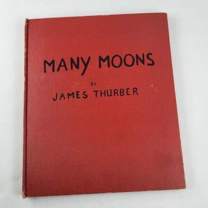 May include: A red hardcover book titled "Many Moons" by James Thurber. The title and author's name are in black font. The book has a textured cover and appears to be vintage.