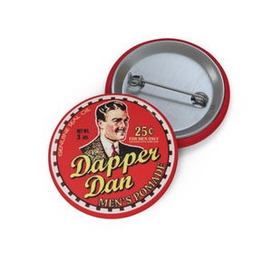 May include: A red button with the text "Dapper Dan Men's Pomade" in yellow script. The button features a vintage illustration of a man and the text "25¢ for men only". The button is a vintage style.