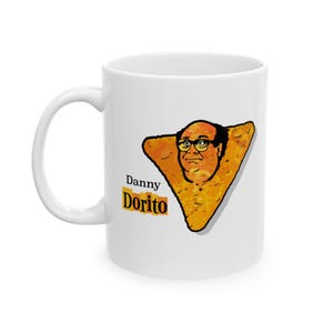 Może przedstawiać: Biały kubek ceramiczny z kreskówkową ilustracją twarzy na żółtym trójkątnym chipsie. Pod chipsem nadrukowany jest tekst "Danny Dorito".