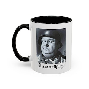 May include: White ceramic coffee mug with a black rim and handle. The mug features a black and white image of a man in a military uniform with the text "I see nothing..." printed below the image.