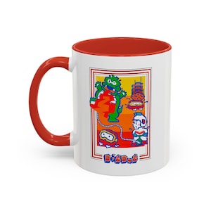 May include: White ceramic coffee mug with a red rim. The mug features a colourful illustration of a video game character, a monster, and a building. The text "Dig Dug" is printed on the bottom of the mug.