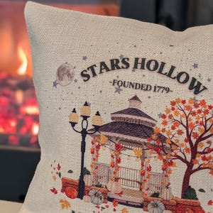 Könnte beinhalten: Cremefarbenes Zierkissen mit einem Aufdruck, der einen Pavillon, Kürbisse und Herbstlaub zeigt. Der Text "STARS HOLLOW - FOUNDED 1779" und "AUTUMN FESTIVAL" sind ebenfalls aufgedruckt. Ein dekorativer Artikel zum Thema Herbst.