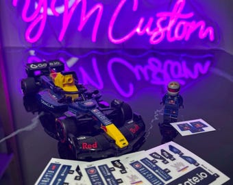 May include: A toy race car in black, red, and yellow with sponsor decals and a Lego figure. The car is positioned in front of a neon sign that reads "yM Custom". Several "Gate.io" stickers are in front of the car.