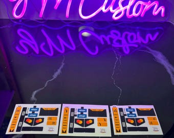 May include: A neon sign in a cursive font that reads "yM Custom" in bright pink. Below the sign are several sticker sheets with orange, black, and white designs. The stickers have the text "always" and "wingo".