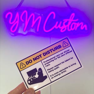 May include: A rectangular white sign with black text and a yellow warning icon. The sign reads DO NOT DISTURB, Professional race car driver preparing for his next official race. Please respect his talent by being quiet. It is even advisable to bring an energy drink. A black graphic of a racing simulator setup is included.