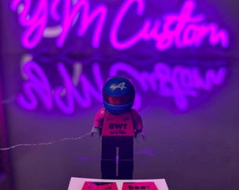 May include: A Lego figurine of a race car driver wearing a blue helmet with the number 44. The figurine is wearing a pink shirt and black trousers. Two pink stickers with the text "BWT" are in front of the figurine. A neon sign in the background reads "YM Custom".