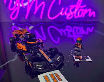 May include: A Lego model of a black and orange race car, with a matching Lego figure and sticker sheet. A neon sign in the background reads "yM Custom" in pink.