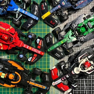 Puede incluir: Una colección de coches de carreras de juguete coloridos, con varios logotipos de patrocinadores y diseños. Los coches son de color rojo, azul, verde y naranja, con neumáticos negros y componentes detallados. La imagen se toma desde arriba.