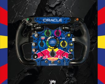 May include: A Formula 1 style racing steering wheel featuring a blue and black digital camouflage pattern, a central Red Bull logo, and various control buttons labeled with text including Oracle, DIF IN, CHRG, and PIT, set against a dark background.