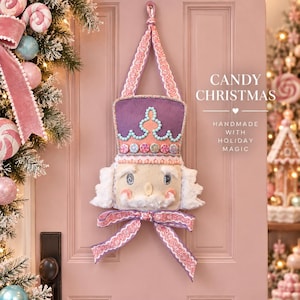 May include: A fabric nutcracker wall hanging featuring a purple hat with teal trim, a beige face with white faux fur hair and beard, and a pink patterned ribbon bow. The text CANDY CHRISTMAS HANDMADE WITH HOLIDAY MAGIC is printed on the right.