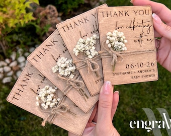May include: Set of five wooden thank you cards with a small bouquet of white flowers tied with twine. The cards are engraved with the text "Thank you for celebrating with us" and the date "06-10-26". The text "Stephanie + Andrew's Baby Shower" is also engraved on the cards.