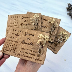 May include: Wooden save the date cards with a rustic design. The cards feature a calendar with the date June 14, 2025, and the names Gemma & Alexander. The cards are decorated with dried flowers and twine.