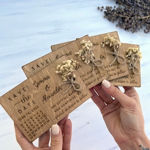 May include: Save the date cards with a rustic wooden design. The cards feature a calendar with the date June 14, 2025, and the names Gemma & Alexander. The cards are decorated with dried flowers and twine.