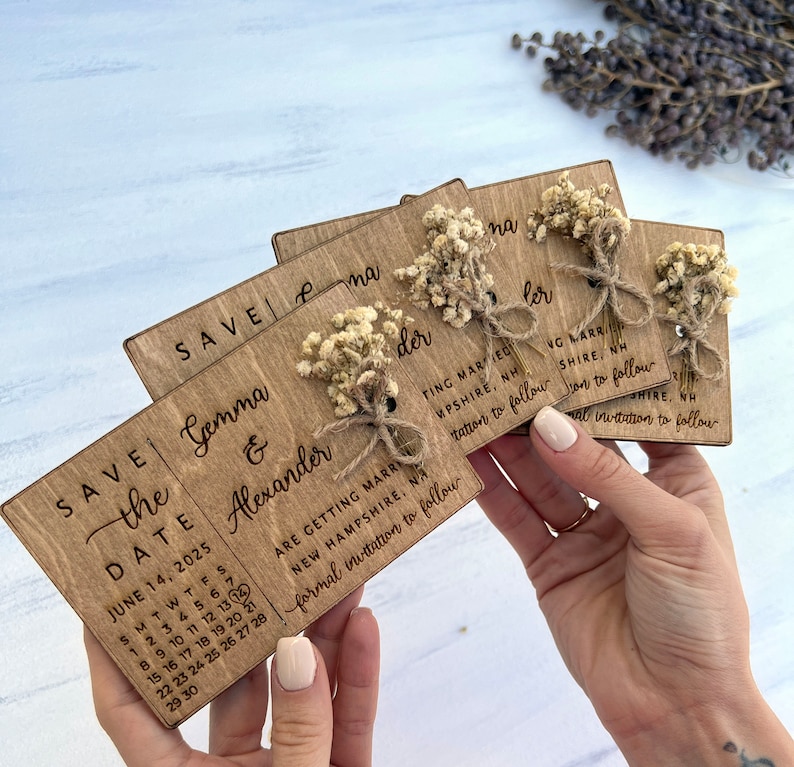 May include: Save the date cards with a rustic wooden design. The cards feature a calendar with the date June 14, 2025, and the names Gemma & Alexander. The cards are decorated with dried flowers and twine.