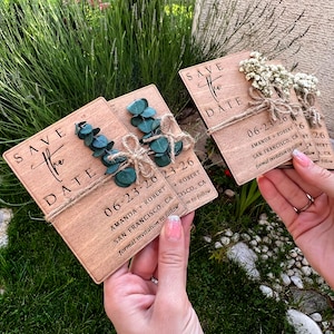 May include: Three wooden save the date cards with greenery and twine accents. The cards are decorated with the text "Save the Date" and the date "06-23-26". The cards also include the names "Amanda + Robert" and the location "San Francisco, CA".