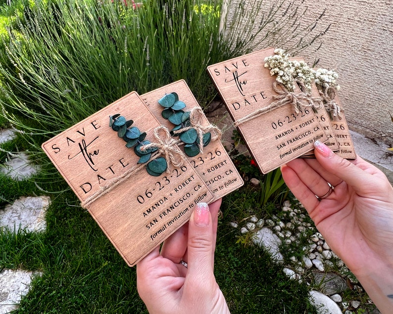 May include: Three wooden save the date cards with greenery and twine accents. The cards are decorated with the text "Save the Date" and the date "06-23-26". The cards also include the names "Amanda + Robert" and the location "San Francisco, CA".