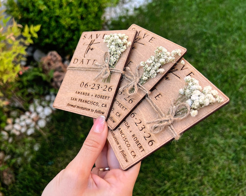 May include: Three wooden save the date cards with a rustic brown finish. Each card has a small bouquet of white baby's breath tied with twine. The text on the cards reads "Save the Date", "06.23.26", "Amanda + Robert", "San Francisco, CA", and "Formal invitation to follow".