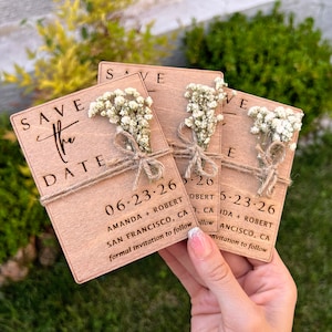 May include: Save the date cards with a rustic wooden design. The cards feature a small bouquet of white flowers and the text "Save the Date" with the date "06.23.26" and the names "Amanda + Robert" and "San Francisco, CA".