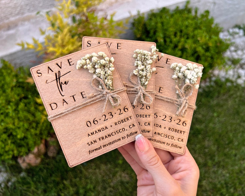 May include: Save the date cards with a rustic wooden design. The cards feature a small bouquet of white flowers and the text "Save the Date" with the date "06.23.26" and the names "Amanda + Robert" and "San Francisco, CA".