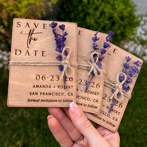 May include: Three wooden save the date cards with lavender sprigs tied with twine. The cards are engraved with the text "Save the Date", the date "06.23.26", the names "Amanda + Robert", the location "San Francisco, CA", and the phrase "formal invitation to follow".