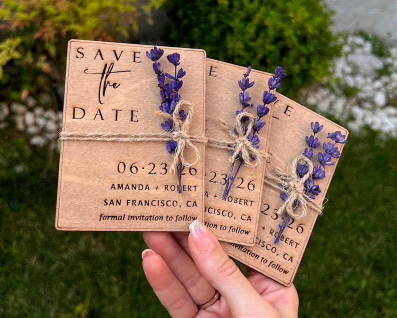 May include: Three wooden save the date cards with lavender sprigs tied with twine. The cards are engraved with the text "Save the Date", the date "06.23.26", the names "Amanda + Robert", the location "San Francisco, CA", and the phrase "formal invitation to follow".