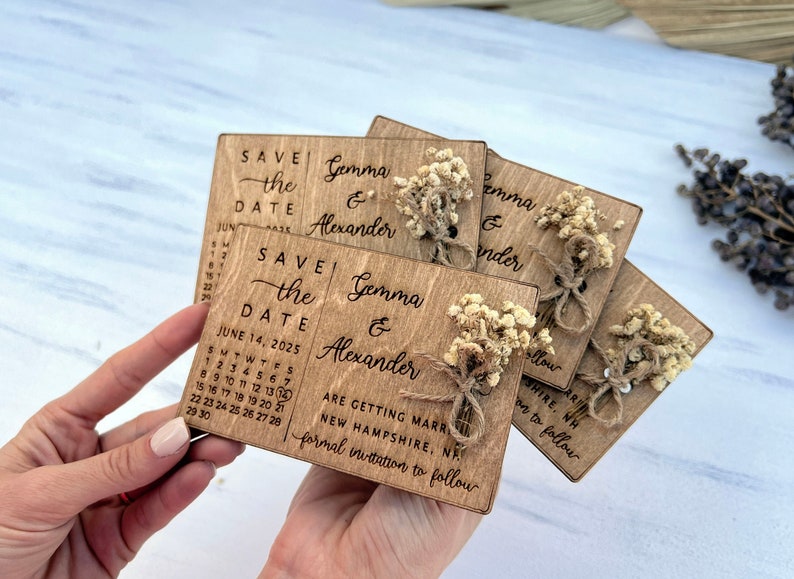 May include: Save the date cards with a rustic wooden design. The cards feature a calendar with the date June 14, 2025, and the names Gemma & Alexander. The cards are decorated with dried flowers and twine.