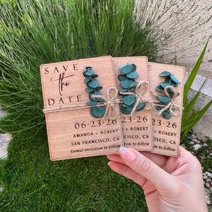 May include: Three wooden save the date cards with greenery and twine. The cards read "Save the Date" with the date "06-23-26" and the names "Amanda + Robert" and the location "San Francisco, CA". The second card reads "23-26" with the names "Robert" and the location "Cisco, CA". The third card reads "23-26" with the names "Robert" and the location "Cisco, CA".