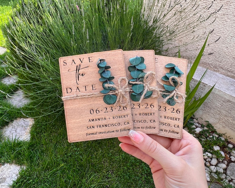 May include: Three wooden save the date cards with greenery and twine. The cards read "Save the Date" with the date "06-23-26" and the names "Amanda + Robert" and the location "San Francisco, CA". The second card reads "23-26" with the names "Robert" and the location "Cisco, CA". The third card reads "23-26" with the names "Robert" and the location "Cisco, CA".