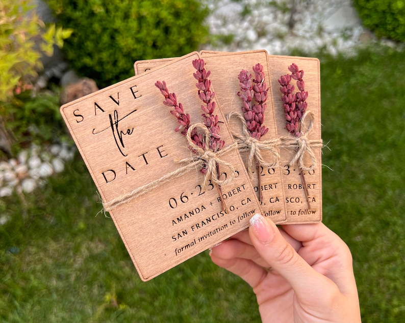 May include: Three wooden save the date cards with a rustic brown finish. Each card has a small bundle of dried lavender tied with twine. The text on the cards reads "Save the Date", "06.23.23", "Amanda + Robert", "San Francisco, CA", and "Formal invitation to follow".