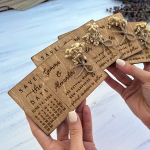 May include: Save the date cards with a rustic wooden design. The cards feature a calendar with the date June 14, 2025, and the names Gemma & Alexander. The cards are decorated with dried flowers and twine.