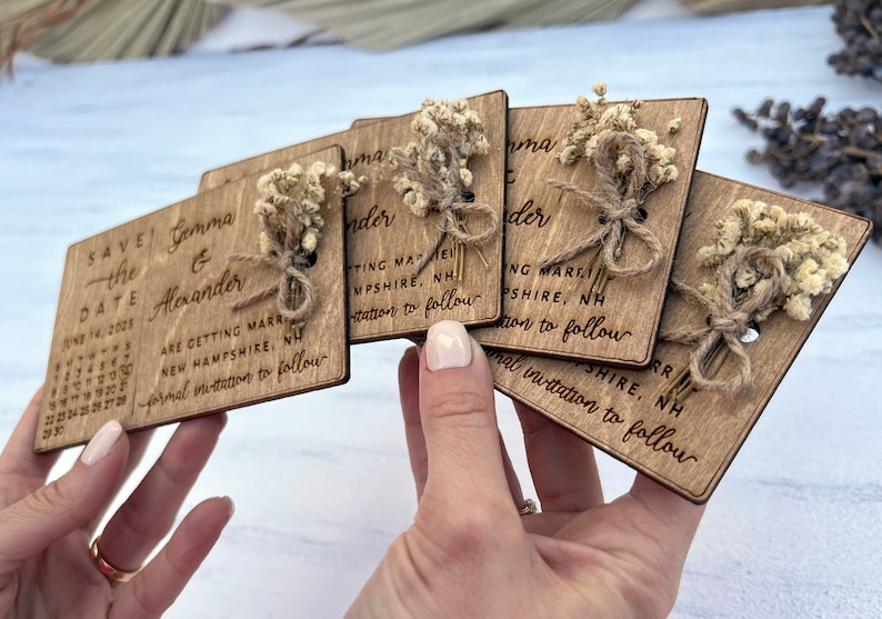 May include: Set of six wooden save the date cards with dried flowers and twine. The cards are engraved with the text "Save the Date", the couple's names, the date, and the location of the wedding. The cards are perfect for a rustic or farmhouse wedding.