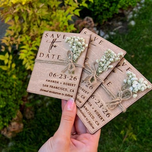 May include: Set of four wooden save the date cards with white flowers and twine. The cards are engraved with the text "Save the Date", the date "06-23-26", the names "Amanda + Robert", and the location "San Francisco, CA".