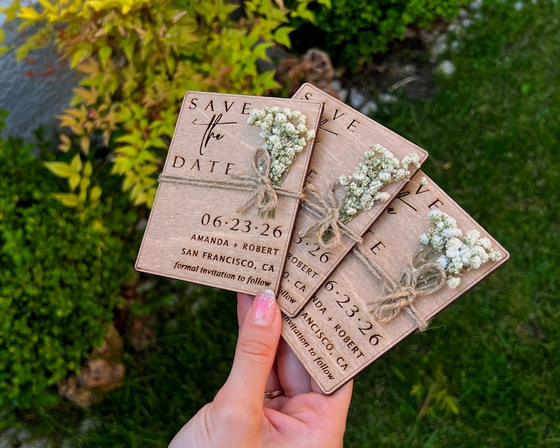 May include: Set of four wooden save the date cards with white flowers and twine. The cards are engraved with the text "Save the Date", the date "06-23-26", the names "Amanda + Robert", and the location "San Francisco, CA".