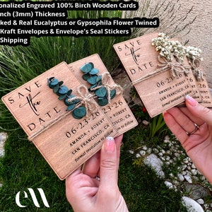 May include: Two wooden save the date cards with the text "Save the Date" and the date "06-23-26" printed on them. The cards are decorated with sprigs of eucalyptus and twine. The cards are held in the hands of a person.