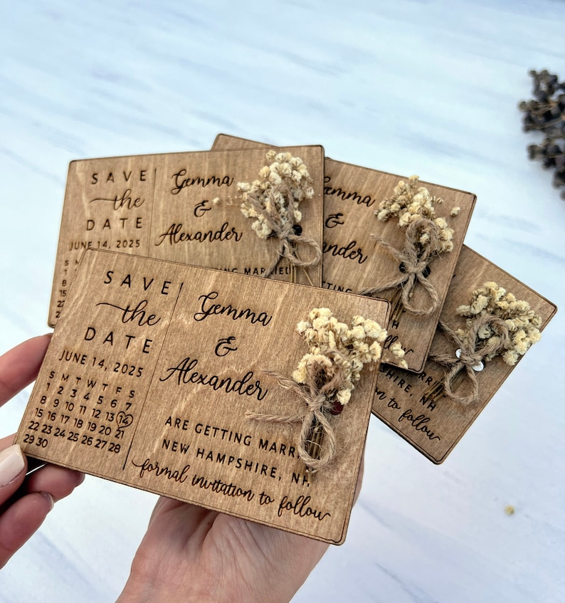 May include: Wooden save the date cards with a rustic design. The cards feature a calendar with the date June 14, 2025, and the names Gemma & Alexander. The cards are decorated with dried flowers and twine.