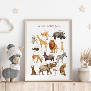 Op de afbeelding: Een witte poster met de titel "WILD ANIMALS" en illustraties van verschillende dieren, waaronder een giraffe, een leeuw, een olifant, een zebra en een leopard. De poster is ingelijst in een licht houten lijst en wordt tentoongesteld op een plank met een knuffeldier en een vaas met gedroogde bloemen.