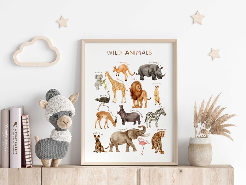 Op de afbeelding: Een witte poster met de titel "WILD ANIMALS" en illustraties van verschillende dieren, waaronder een giraffe, een leeuw, een olifant, een zebra en een leopard. De poster is ingelijst in een licht houten lijst en wordt tentoongesteld op een plank met een knuffeldier en een vaas met gedroogde bloemen.