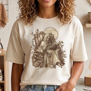 Puede incluir: Una camiseta de manga corta color crema con un estampado gráfico marrón de un esqueleto con capucha sentado en una silla leyendo un libro, rodeado de elementos florales y una luna llena. La camiseta se combina con unos vaqueros azul claro.
