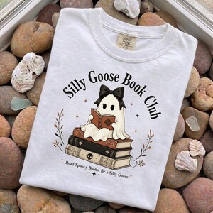 Puede incluir: Una camiseta blanca con el gráfico de un fantasma con un lazo negro, leyendo un libro mientras está sentado sobre una pila de tres libros. El texto dice Silly Goose Book Club y Read Spooky Books, Be a Silly Goose. La camiseta tiene cuello redondo.