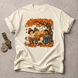 Puede incluir: Una camiseta de color crema con una ilustración gráfica de cuatro niños y un perro pequeño sentados en un banco bajo un árbol de otoño con hojas naranjas. Hay calabazas esparcidas por el suelo. Un reloj de pulsera está a la izquierda.