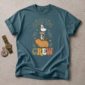 May include: A teal short-sleeved t-shirt featuring a graphic of a capybara, a raccoon with a backpack, and a duck with a pink bow stacked vertically. The text reads "it's a GOOD DAY CREW" in a retro font, surrounded by small hearts and leaf sprigs.